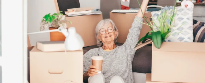 Senior woman relaxing among moving boxes with a coffee—downsizing tips.