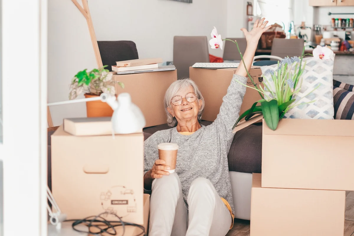 downsizing-tips-senior-woman-moving-boxes Senior woman relaxing among moving boxes with a coffee—downsizing tips.
